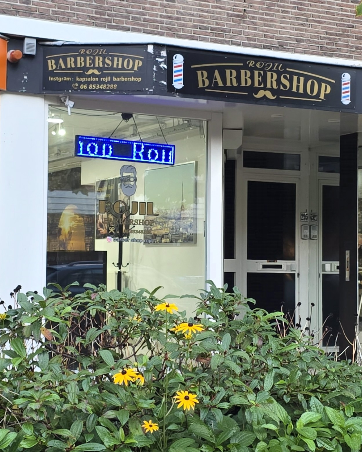 De zaak vanaf de stoep: het zwarte bord met Barbershop, de etalage en de bloembak ervoor